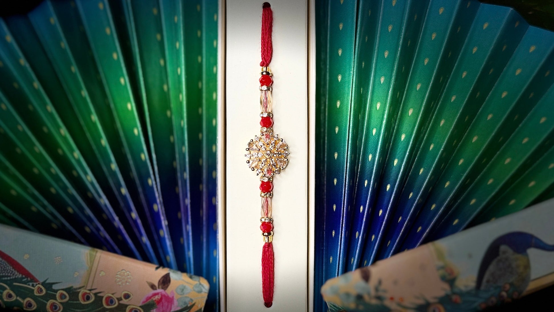 Decorative hand fan with peacock design and a red rakhi on a black background